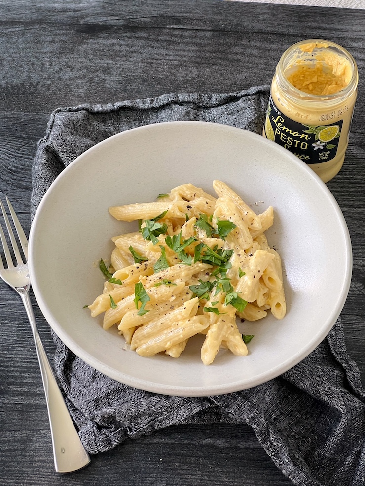 Penne with Trader Joe's Lemon Pesto served in aw hite bowl garnished with parsley. The pesto jar is in the background, a fork on the left of the bowl.