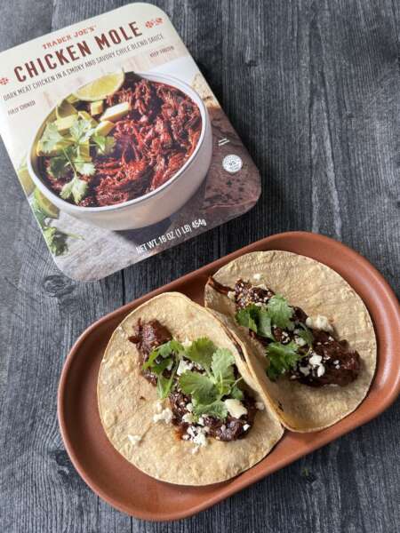 Trader Joe's Chicken Mole box next to an oval plate with two mole tacos topped cotija, sesame seeds and cilantro.