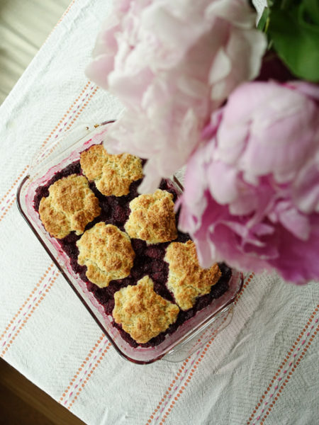 A glass dish of berry cobbler is shot from above with peonies peeking into the frame.