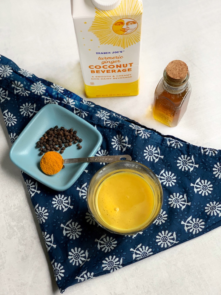 Trader Joe's Turmeric Ginger Coconut Beverage in a small glass is set next to a small dish with a teaspoon of turmeric and black peppercorns.