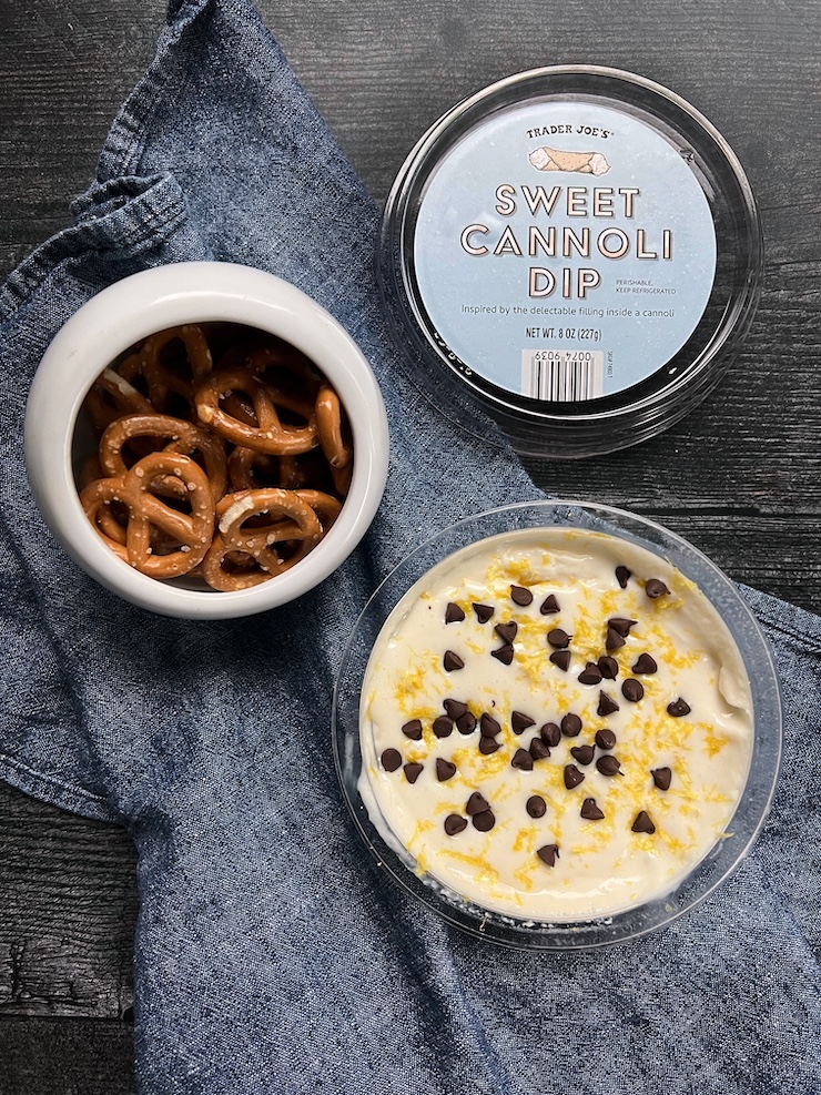 An open pint container of Trader Joe's Sweet Cannoli dip garnished with grated lemon zest and mini chocolate chips. Mini twist pretzels are situation to it left and the lid displaying the name "Sweet Cannoli Dip" is above the tub.