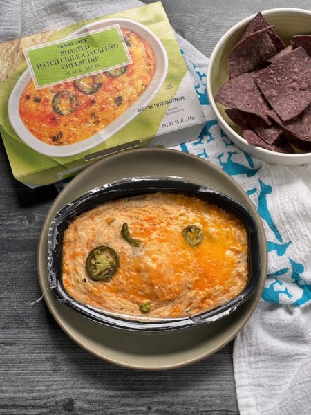 Trader JOe's Roasted hatch Chile & Jalapeno Cheese Dip sleeve & dip arranged with a bowl of blue corn tortilla chips on a weathered wood background.