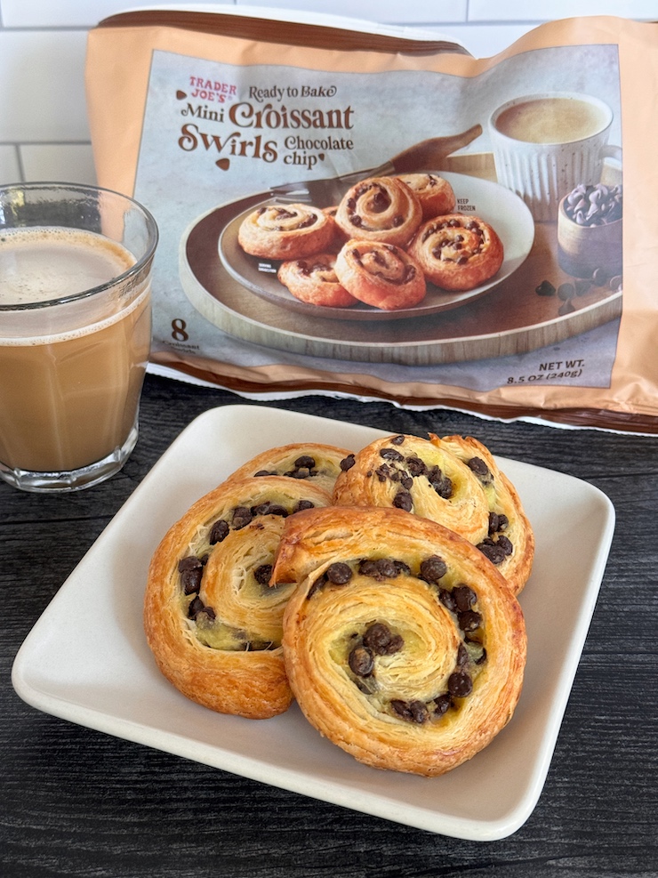 Trader Joe's Chocolate Chip Mini Croissant Swirls stacked on a white plate with a cup of coffee. The package is in the background.