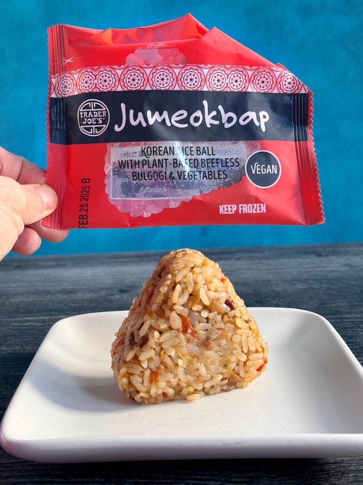 Trader Joe's Jumeokbap, a triangular Korean rice ball sitting vertically on a white plate. It's package is held up above it.