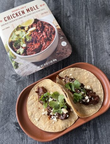 Trader Joe's Chicken Mole box beside an oval plate with two chicken mole tacos on corn tortillas topped with cotija and cilantro.